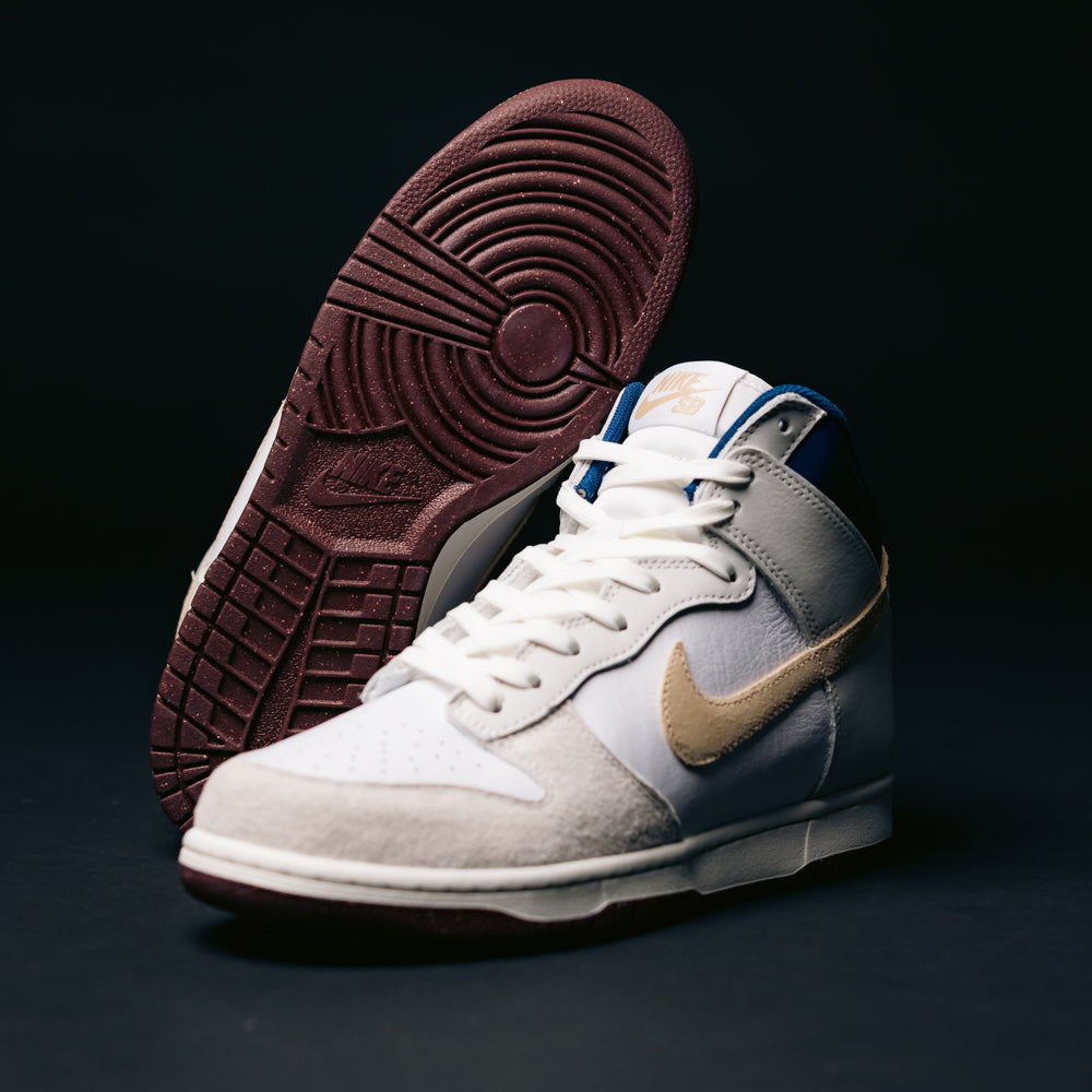 The Ivory Sand Blue Void Dunk High Pro x Beyond IU4491100 Nike SB Skateboarding Shoes, featuring white, tan, and gray panels with a light brown suede swoosh and maroon soles, are displayed against a dark background—one shoe standing upright while the other lies on its side.