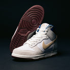The Ivory Sand Blue Void Dunk High Pro x Beyond IU4491100 Nike SB Skateboarding Shoes, featuring white, tan, and gray panels with a light brown suede swoosh and maroon soles, are displayed against a dark background—one shoe standing upright while the other lies on its side.