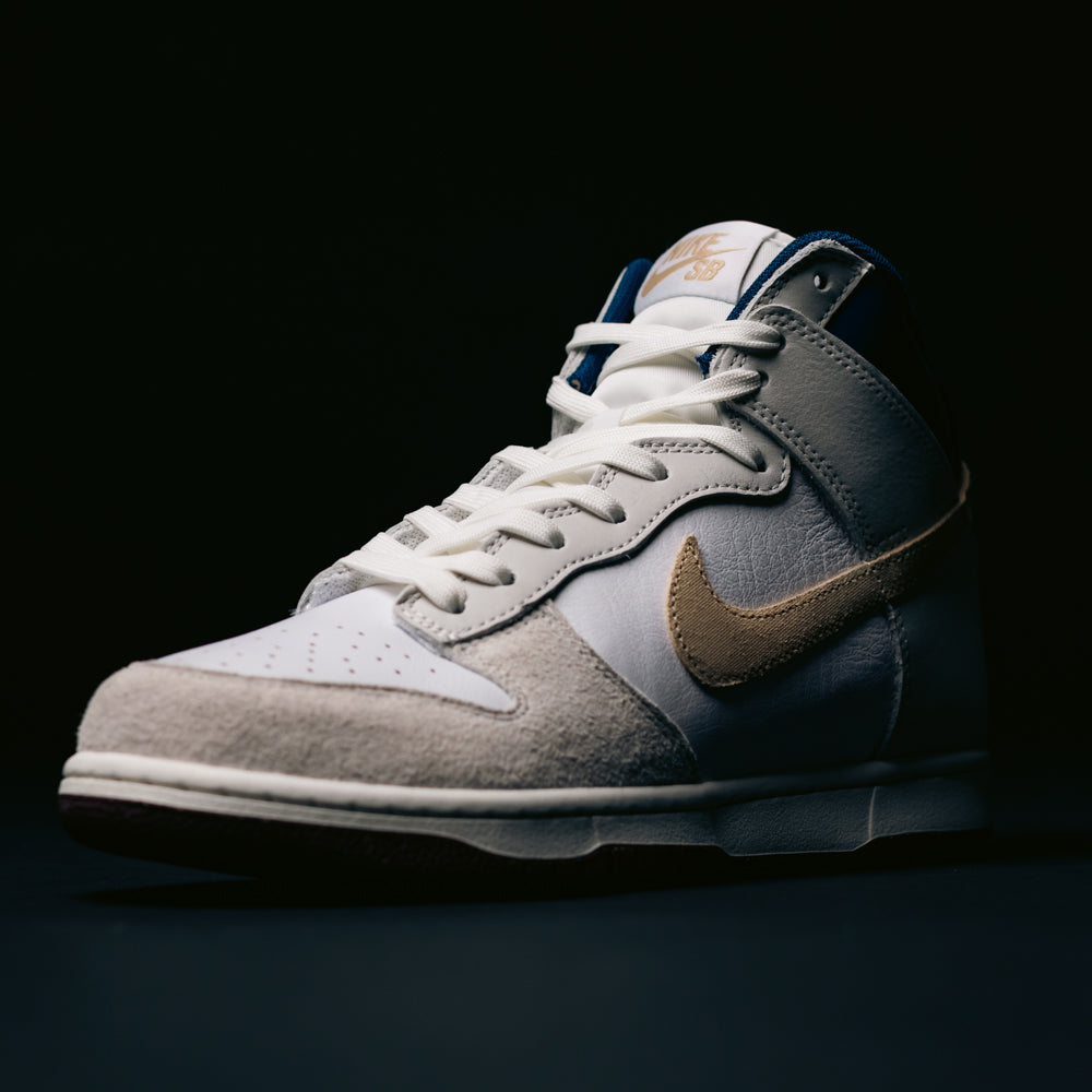 An Ivory Sand and Blue Void Dunk High Pro x Beyond Nike SB skateboarding shoe (model IU4491100) is displayed against a dark background, featuring a gold Nike swoosh and dramatic side lighting.