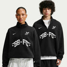 Two people wear imported Black IM5463010 Skate 1/2 Zip Fleece Crew Nike SB Skateboarding sweatshirts with white SB-FG text, stars, and winged logos. The left has hair tied back; the right has curly hair and layered necklaces. Machine wash recommended.