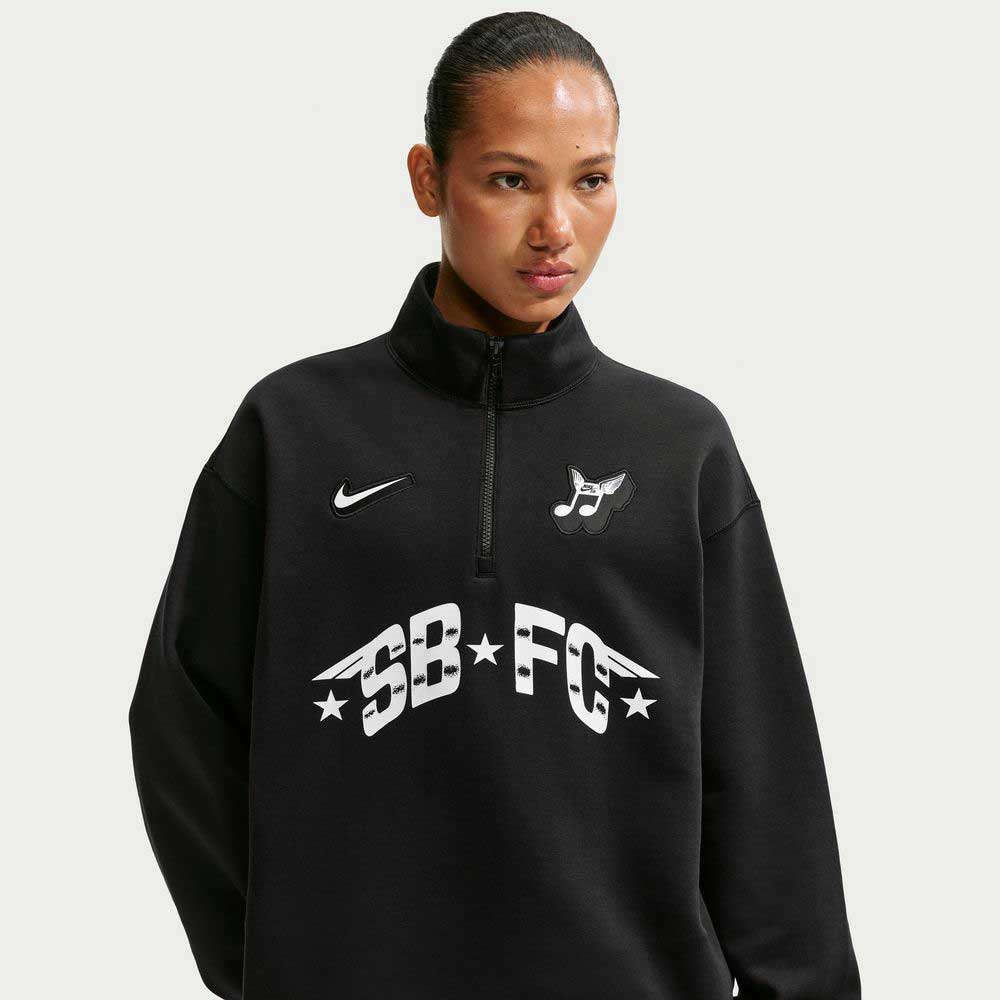 Wearing the Black IM5463010 Skate 1/2 Zip Fleece Crew from Nike SB Skateboarding, featuring white logos and text, a person stands against a plain light background. Machine washable and easy to care for.