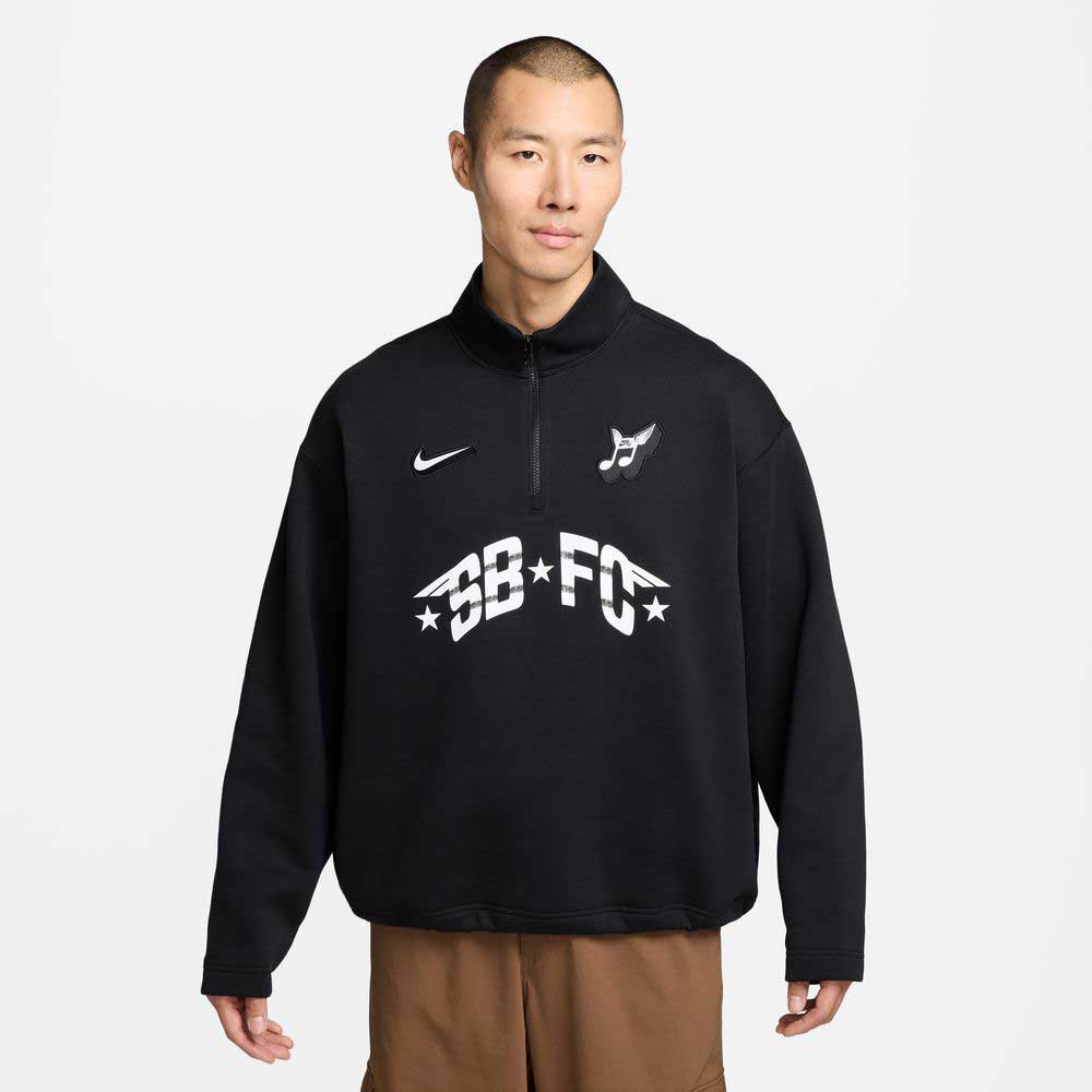 A person with short hair wears a black Nike SB Skateboarding IM5463010 1/2 Zip Fleece Crew with white logos and BB FC text, paired with brown pants, standing against a plain light background. The sweatshirt is machine washable.