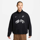 A person with short hair wears a black Nike SB Skateboarding IM5463010 1/2 Zip Fleece Crew with white logos and BB FC text, paired with brown pants, standing against a plain light background. The sweatshirt is machine washable.