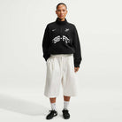 A person stands facing forward, wearing the Black IM5463010 Skate 1/2 Zip Fleece Crew from Nike SB Skateboarding with loose white shorts, white socks, and black sneakers against a plain light background. Machine washable for easy care.