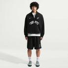 A young person with curly hair stands against a white background wearing the imported black Nike SB Skateboarding IM5463010 Skate 1/2 Zip Fleece Crew, styled with black shorts, white socks, gray sneakers, and featuring white front graphics and easy machine wash care.