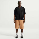 A person stands against a plain white background, wearing the Black Jersey Tee IF1302010 T-Shirt Nike SB Skateboarding Tee, tan knee-length shorts, white socks, black sneakers, with braided short hair and a simple necklace.