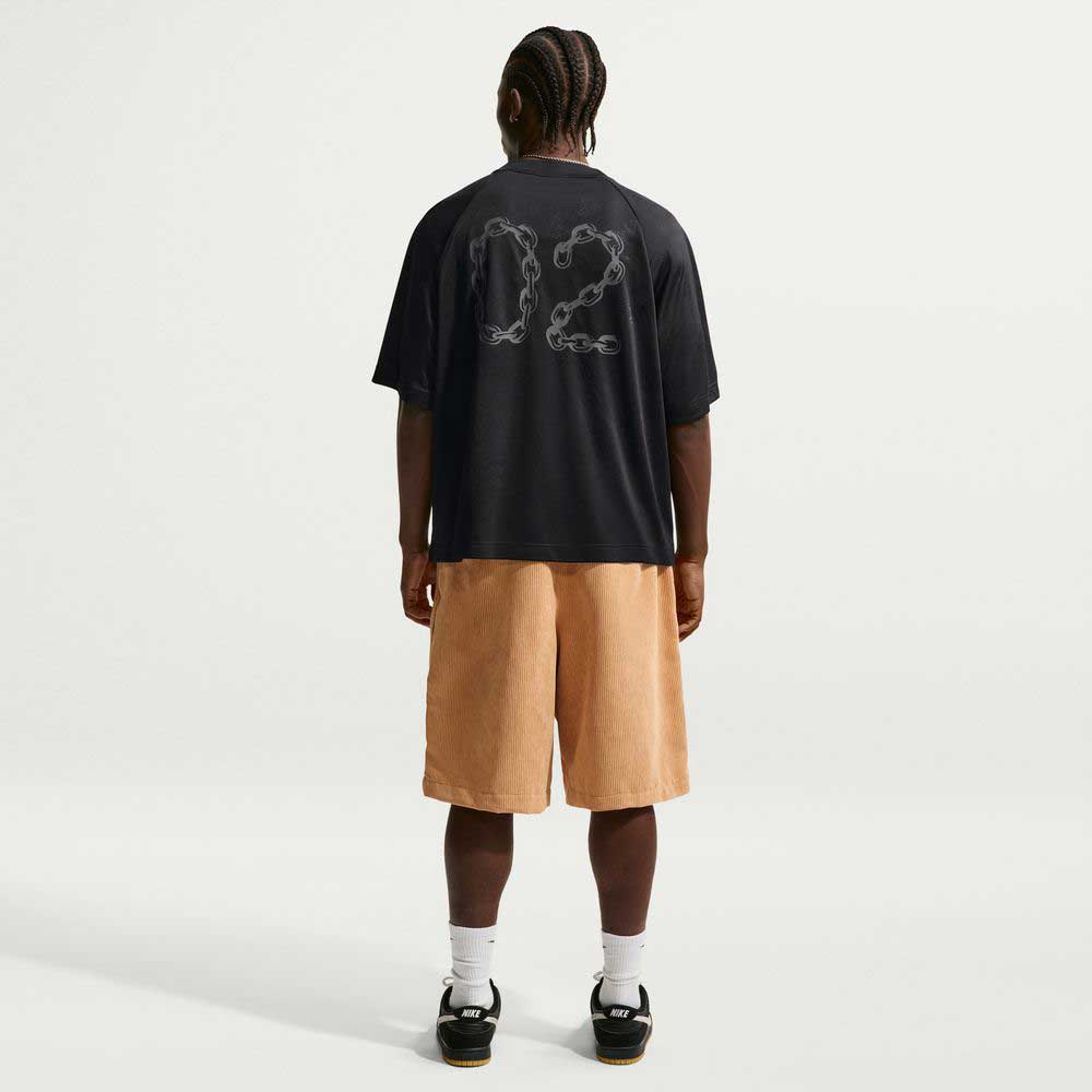 A person stands facing away, wearing the Black Jersey Tee IF1302010 T-Shirt Nike SB Skateboarding Tee with '02' on the back, tan knee-length shorts, white socks, and black sneakers, against a plain white background. The tee offers sweat-wicking comfort.