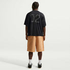 A person stands facing away, wearing the Black Jersey Tee IF1302010 T-Shirt Nike SB Skateboarding Tee with '02' on the back, tan knee-length shorts, white socks, and black sneakers, against a plain white background. The tee offers sweat-wicking comfort.