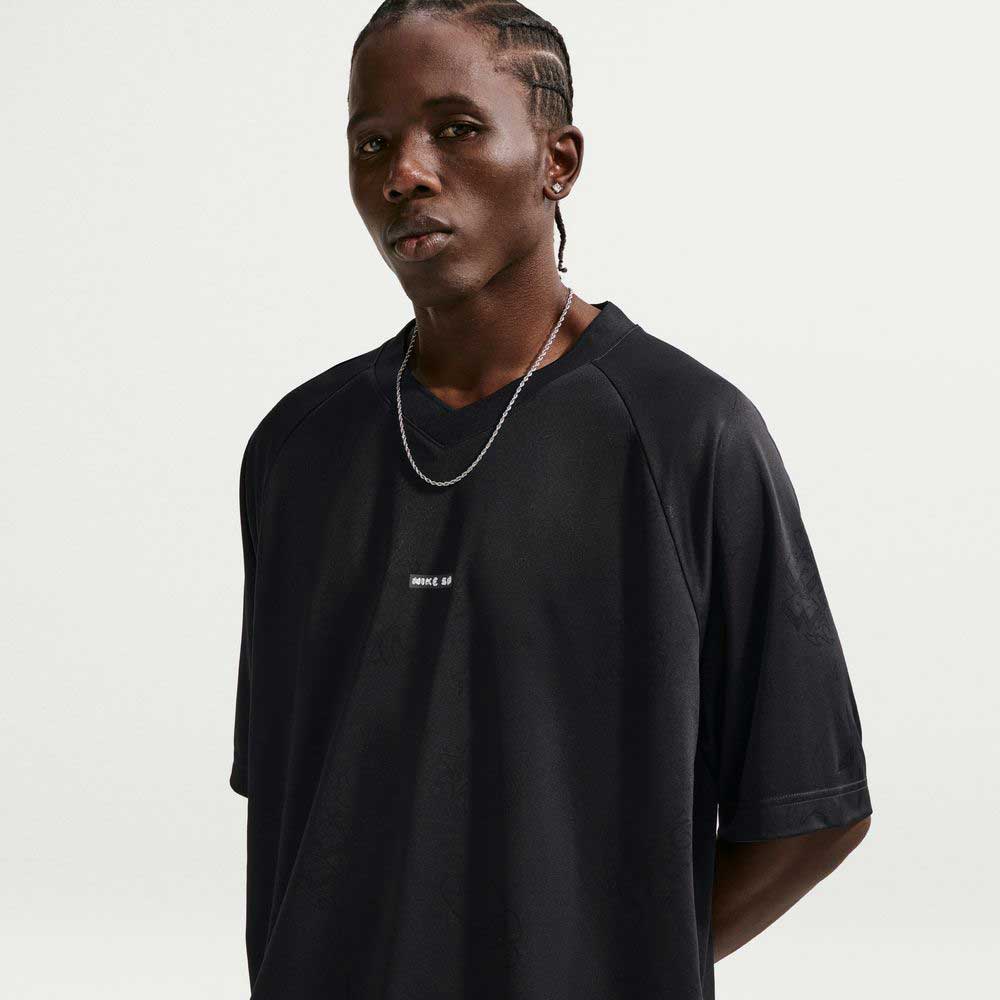 A person with braided hair wears a Black Jersey Tee IF1302010 T-Shirt Nike SB Skateboarding Tee and a silver chain necklace, standing against a plain light background and looking slightly to the side.