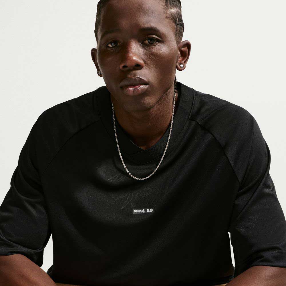 A person with braided hair, wearing a Black Jersey Tee IF1302010 T-Shirt Nike SB Skateboarding Tee and a silver chain necklace, sits against a plain light background, looking directly at the camera with a neutral expression.