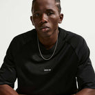 A person with braided hair, wearing a Black Jersey Tee IF1302010 T-Shirt Nike SB Skateboarding Tee and a silver chain necklace, sits against a plain light background, looking directly at the camera with a neutral expression.