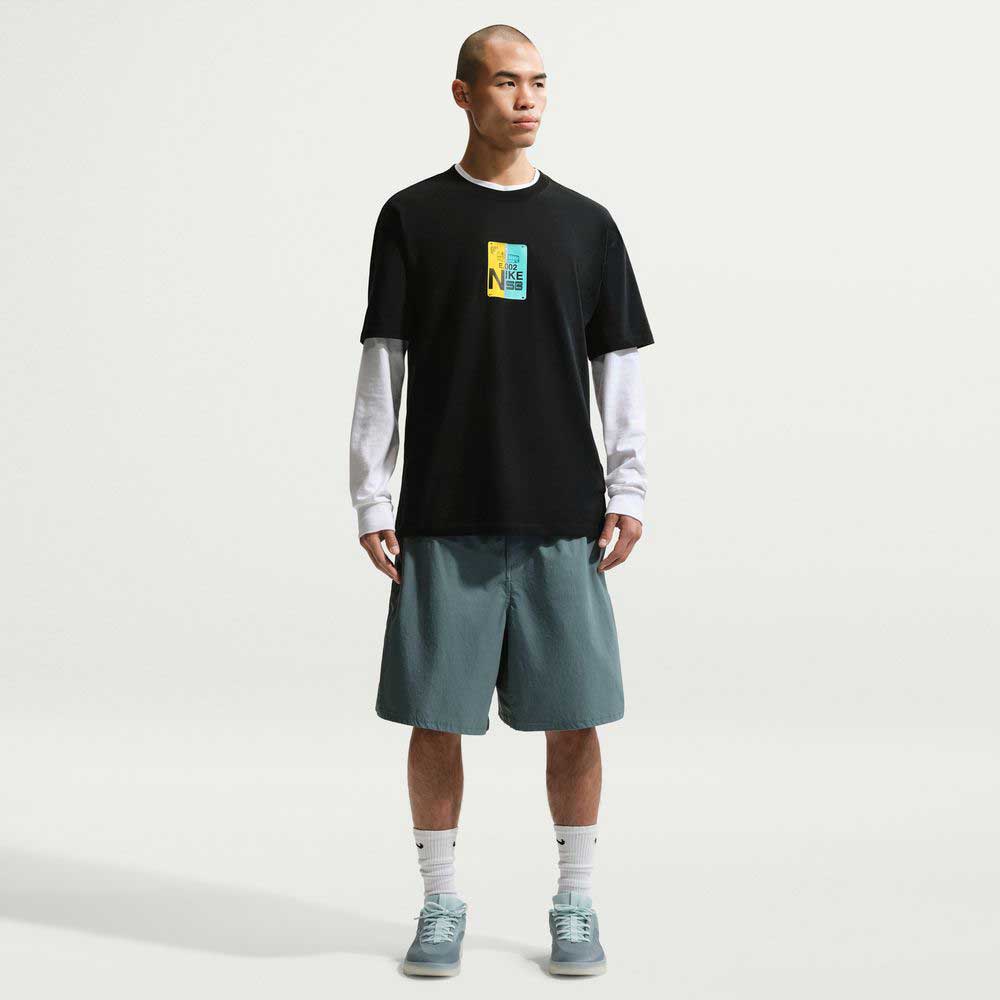 A man faces forward in the Black SF Muni Bus Pass Tee IU1265010 T-Shirt Nike SB Skateboarding Tee, layered over a white long-sleeve, with teal shorts, white socks, and teal sneakers—style and comfort against a plain white background.