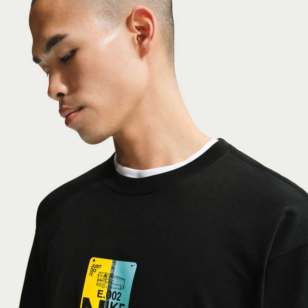 A young man with a buzz cut wears the Black SF Muni Bus Pass Tee IU1265010 Nike SB Skateboarding T-Shirt over a white shirt, looking down against a plain light background.