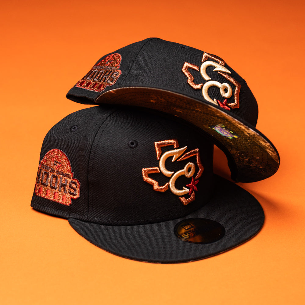 Two black Hooked in Camo Corpus Christi Hooks New Era Hat 5950 Fitted snapbacks with orange and red embroidered logos and patches are displayed on an orange background. One hat is stacked atop the other, revealing a patterned underbrim featuring a subtle Black Camo design reminiscent of the classic 5950 Fitted style.