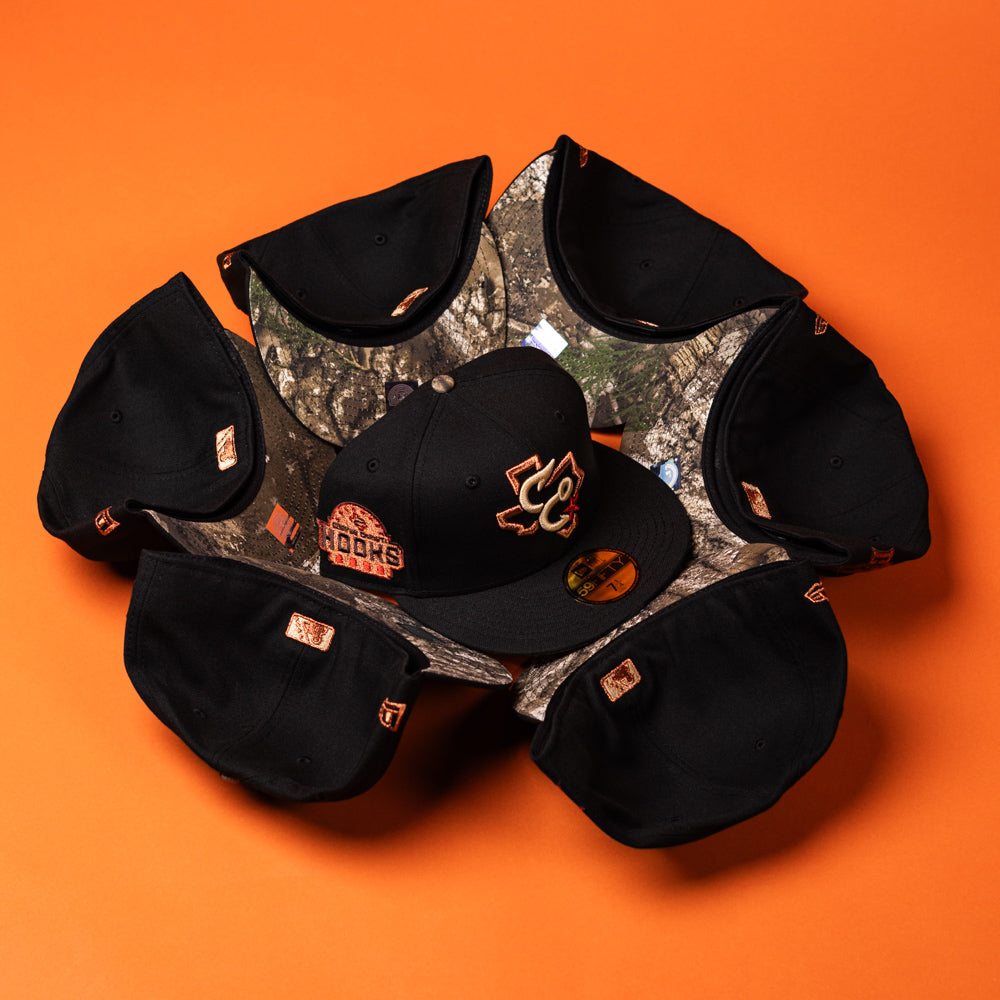 Eight black baseball caps are arranged in a circle on an orange background with their brims facing outward, featuring the Hooked in Camo Corpus Christi Hooks New Era Hat 5950 Fitted at the center, distinguished by its Black Camo brim and Corpus Christi Hooks team logo on the front.