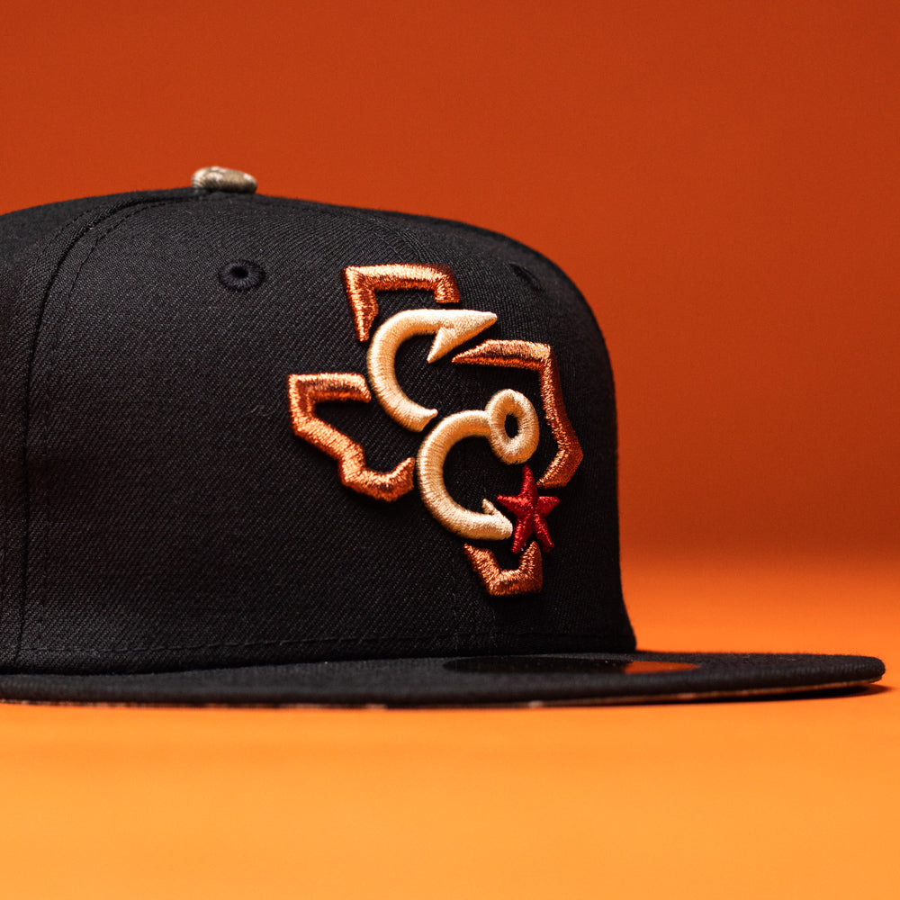 The Hooked in Camo Corpus Christi Hooks New Era Hat 5950 Fitted is a black fitted cap featuring an embroidered outline of Texas with orange and tan C shapes and a red star on the front, displayed against an orange background.