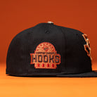The Hooked in Camo Corpus Christi Hooks New Era Hat 5950 Fitted is a black fitted cap with a red and orange embroidered Corpus Christi Hooks logo on the side, displayed against an orange background.