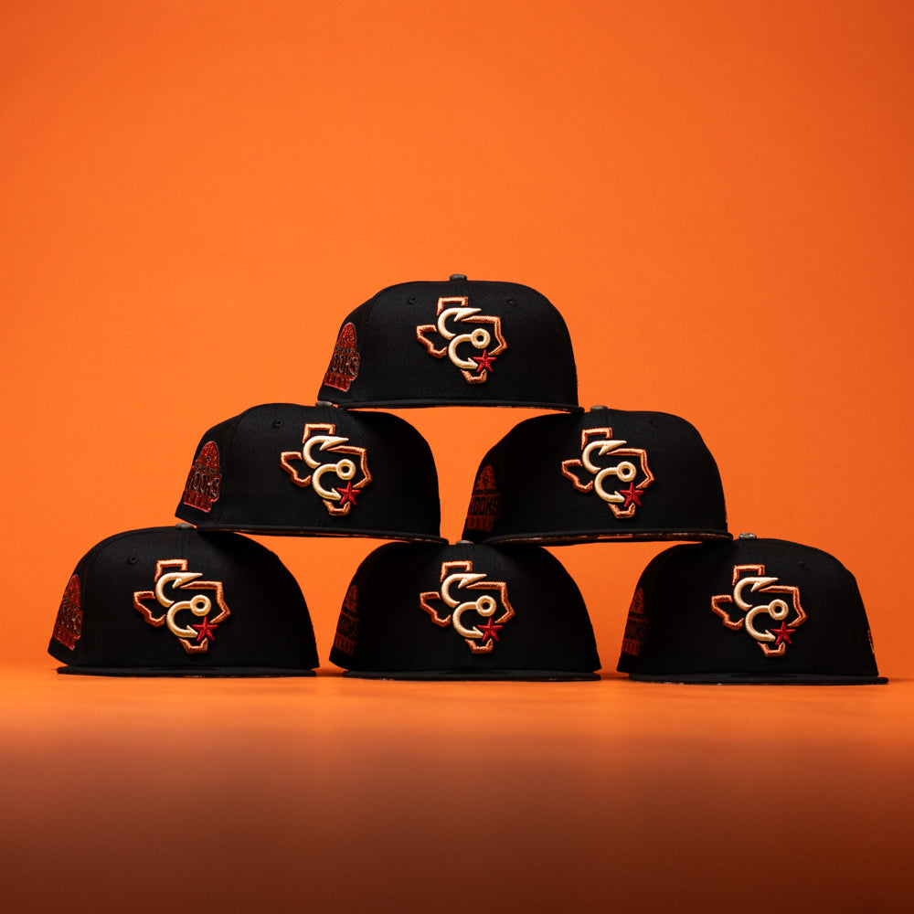 Six black caps—including the Hooked in Camo Corpus Christi Hooks New Era Hat 5950 Fitted, which features orange and white embroidered logos—are stacked in a pyramid shape against a solid orange background.