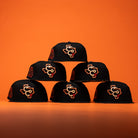 Six black caps—including the Hooked in Camo Corpus Christi Hooks New Era Hat 5950 Fitted, which features orange and white embroidered logos—are stacked in a pyramid shape against a solid orange background.