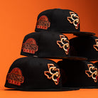 Six Hooked in Camo Corpus Christi Hooks New Era Hat 5950 Fitted caps are stacked in two uneven columns against an orange background. Each black cap features bold orange and red embroidered logos—including a Texas state outline and the word HOOKS—showcasing the distinctive style of the Corpus Christi Hooks.