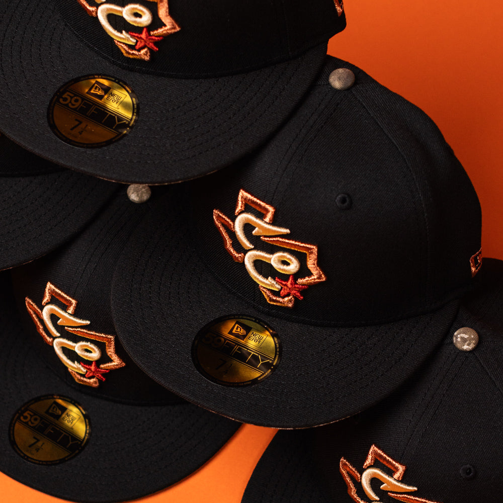 Hooked in Camo Corpus Christi Hooks New Era Hat 5950 Fitted caps, featuring a black camo design with an orange and white embroidered Corpus Christi Hooks logo and gold New Era 59FIFTY stickers on the brims, are stacked on an orange background.