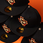 Hooked in Camo Corpus Christi Hooks New Era Hat 5950 Fitted caps, featuring a black camo design with an orange and white embroidered Corpus Christi Hooks logo and gold New Era 59FIFTY stickers on the brims, are stacked on an orange background.