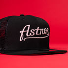 The Black Scarlet Trucker Houston Cougars New Era Hat 5950 Fitted features black mesh and script embroidery, perfect for fans seeking classic style with a fitted trucker look. Displayed on a bold red background.