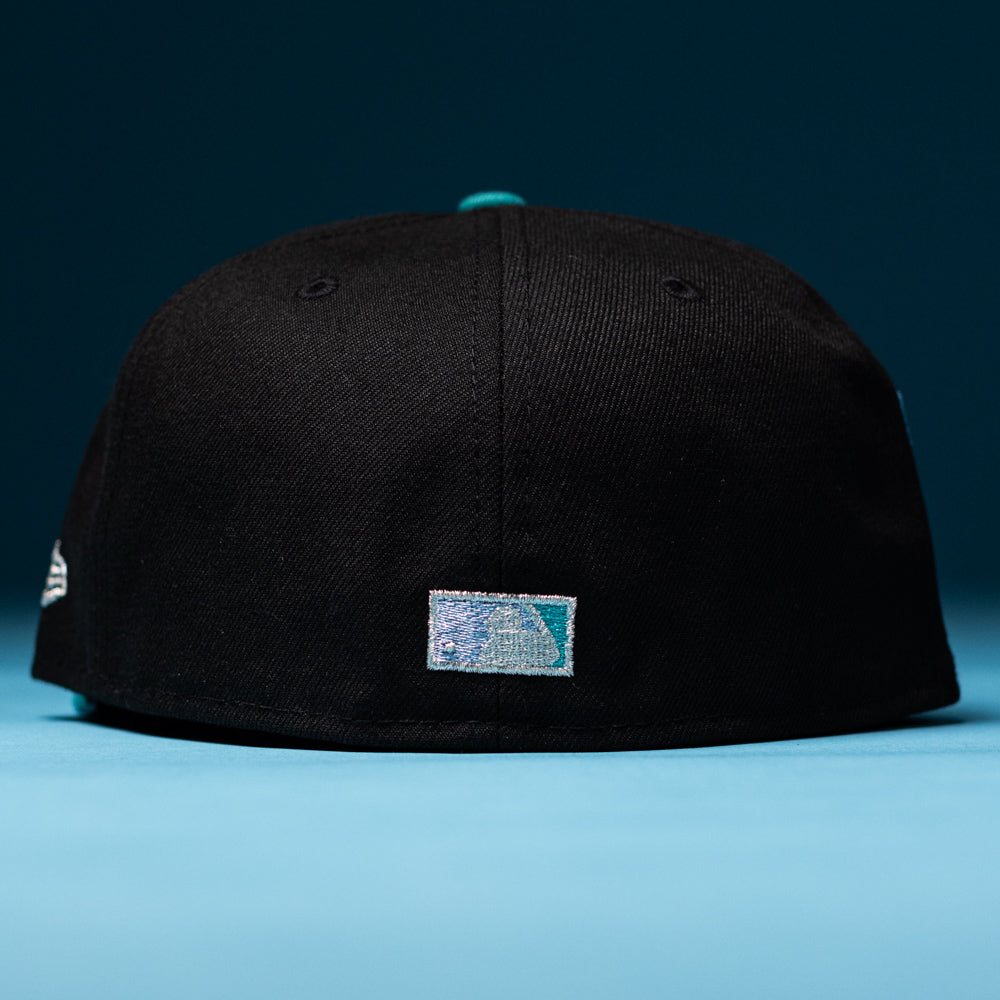 A Silver Lining Black Teal 2000 Houston Astros New Era Hat 5950 Fitted is displayed with its back facing the camera, featuring a small rectangular patch with a blue gradient and white design, set against a blue background.