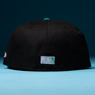 A Silver Lining Black Teal 2000 Houston Astros New Era Hat 5950 Fitted is displayed with its back facing the camera, featuring a small rectangular patch with a blue gradient and white design, set against a blue background.