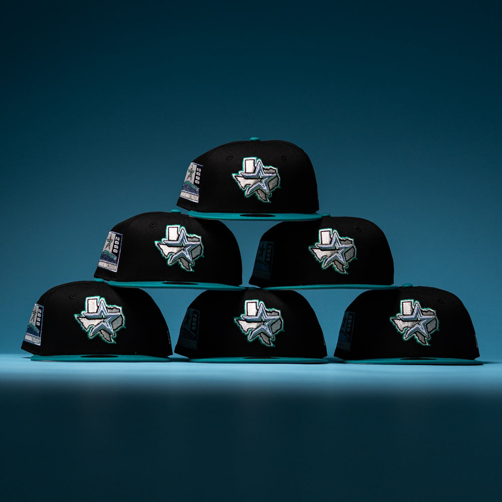 Six Silver Lining Black Teal 2000 Houston Astros New Era 5950 fitted hats are stacked in a pyramid formation, each featuring the signature star and state outline logo on the front. The bold black and teal colorway stands out strikingly against a blue gradient background.