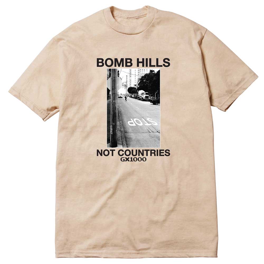 Sand GX1000 tee featuring a black and white photo of a street with STOP painted on the road. Above the image, “Bomb Hills Not Countries” appears in bold black font; below: NOT COUNTRIES GX1000. Product name: Bomb Hills Not Countries Sand GX1000 Tee Black Font.