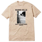 Sand GX1000 tee featuring a black and white photo of a street with STOP painted on the road. Above the image, “Bomb Hills Not Countries” appears in bold black font; below: NOT COUNTRIES GX1000. Product name: Bomb Hills Not Countries Sand GX1000 Tee Black Font.