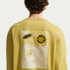 A person is shown from behind wearing the Buff Gold Crew Fleece Nike SB II4440720, featuring a graphic on the back with a large eye, desert landscape, and overlapping circular Nike SB logos.