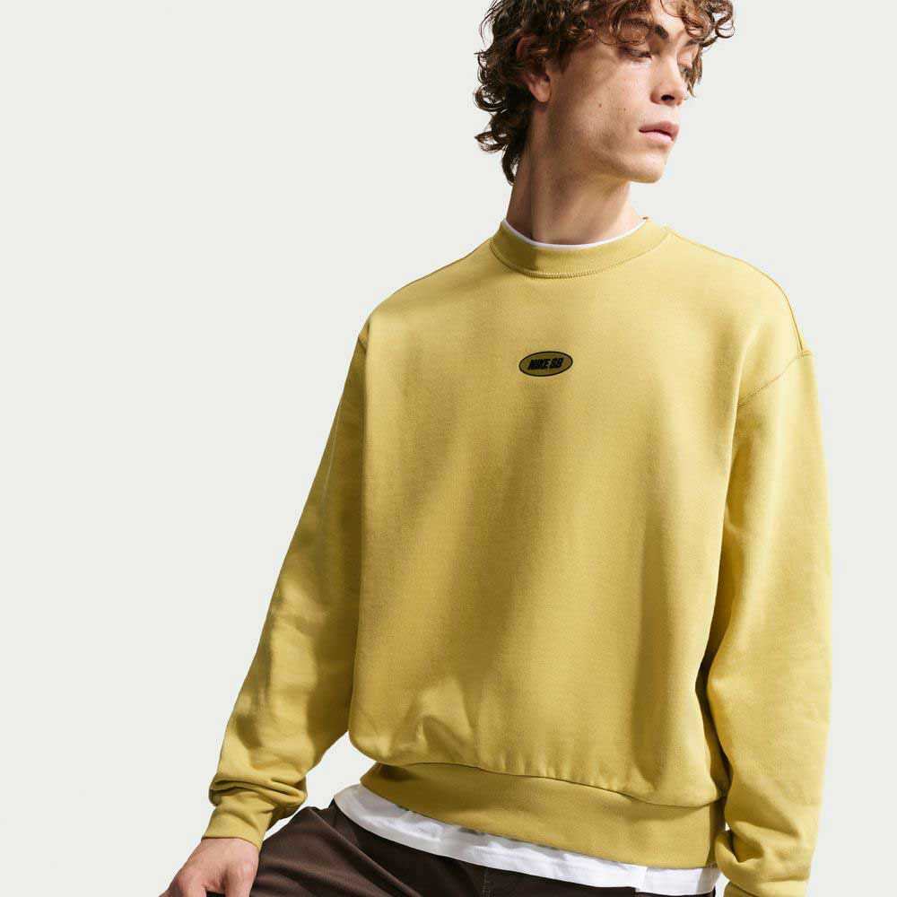 A young person with curly hair wears the Buff Gold Crew Fleece Nike SB II4440720 over a white shirt, looking to the side against a plain light background.