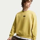 A young person with curly hair wears the Buff Gold Crew Fleece Nike SB II4440720 over a white shirt, looking to the side against a plain light background.