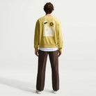 A person faces away, wearing a Buff Gold Crew Fleece Nike SB II4440720 with graphic back designs, brown pants, and white shoes, against a plain white background.