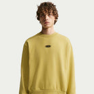 A young man with curly hair wears the Buff Gold Crew Fleece Nike SB II4440720, featuring a small black and white oval logo on the chest, standing against a plain white background.