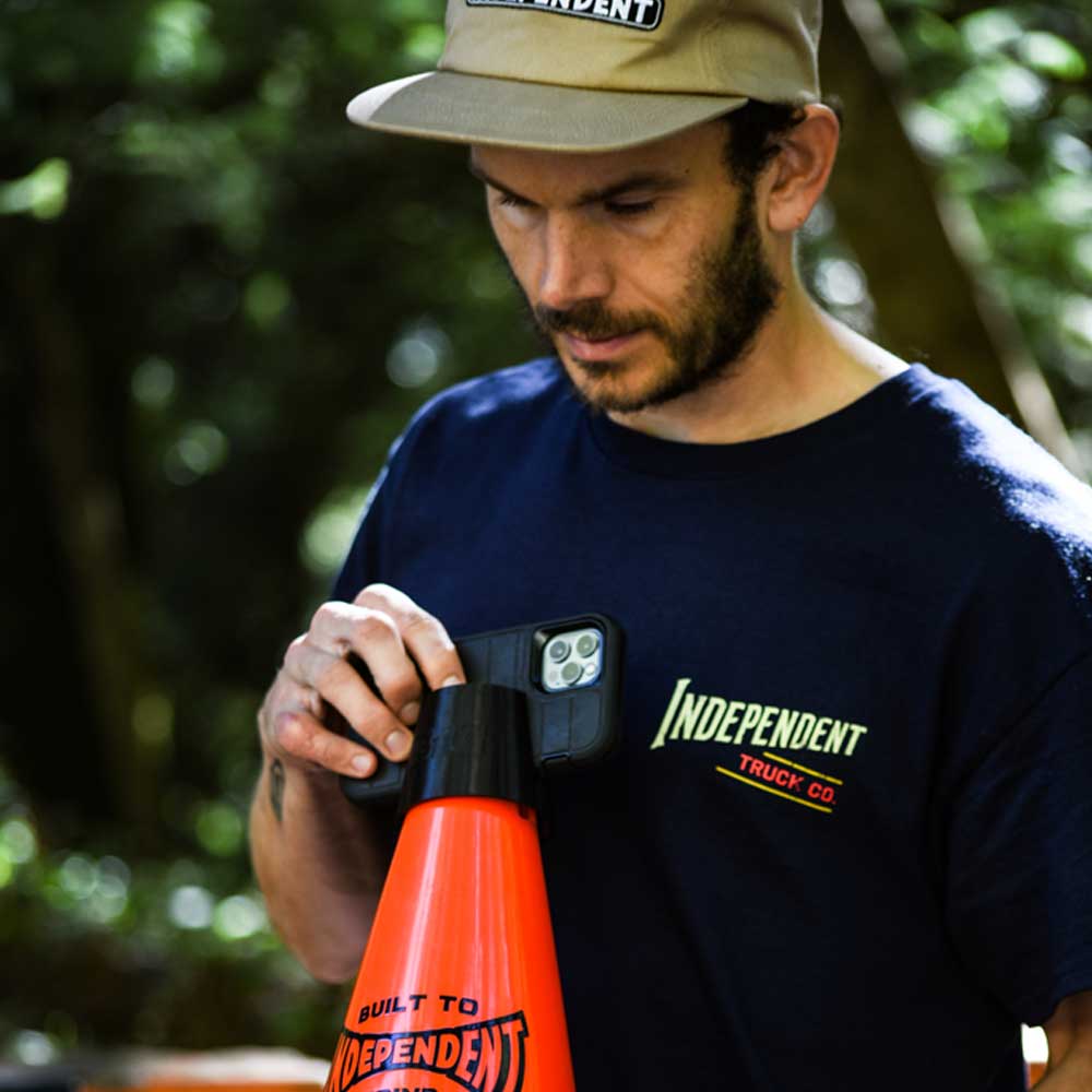 A man outdoors, surrounded by greenery and embodying the Built to Grind spirit of Independent Trucks, holds the Built to Grind Independent Phone Cone while wearing an Independent Truck Co shirt and cap.