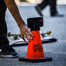 A person places a Built to Grind Independent Phone Cone—shaped like an orange traffic cone and labeled Independent—by a skateboard on a paved surface, turning it into the perfect impromptu phone stand and proving that Independent truly is Built to Grind in every situation.