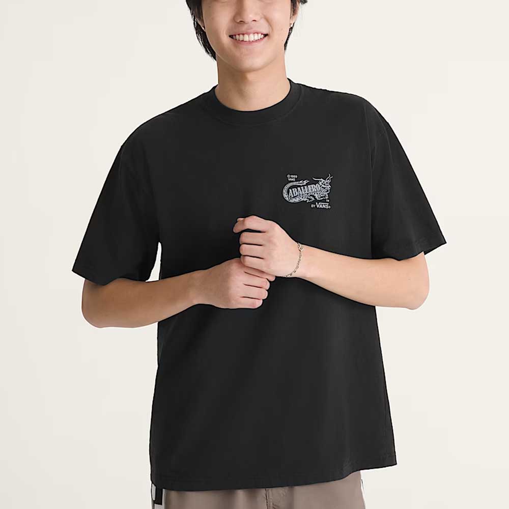 A person wears the Caballero VN000XHBBLK1 Black Short Sleeve Tee by Vans Skateboarding, featuring a small white dragon graphic and text on the chest, standing against a plain light background with hands clasped and smiling.