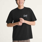 A person wears the Caballero VN000XHBBLK1 Black Short Sleeve Tee by Vans Skateboarding, featuring a small white dragon graphic and text on the chest, standing against a plain light background with hands clasped and smiling.