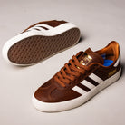 The Brown White Gold Gazelle ADV HQ7547 Adidas Skateboarding Suede Shoes feature a skate-inspired design with white soles and stripes; one shoe stands upright showing the side, while the other lies on its side revealing the honeycomb-patterned sole.