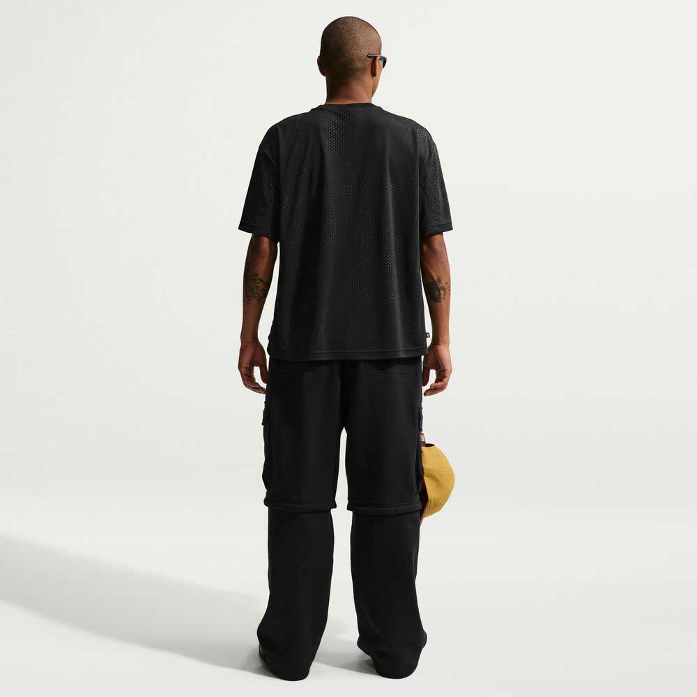 A person with short hair, sunglasses, a black short-sleeve shirt, Ishod Cargo Pant Sweatpant Black IF0659010 Nike SB, and holding a tan cap stands facing away against a plain white background.