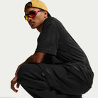 A young man squats in a relaxed pose, wearing Nike SB Ishod Cargo Pant Sweatpant in black, paired with a yellow backwards cap and reflective sunglasses. A black Nike logo is visible. The background is plain and light-colored.