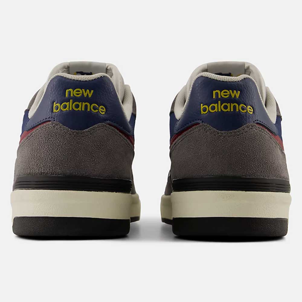 A rear view of the Castlerock Navy Jamie Foy Cup 306 CYZ New Balance Numeric Suede Skateboarding Shoes showcases their mainly grey suede upper with navy and red accents, highlighted by yellow "New Balance" text on the heel tabs against a light background. Inspired by pro skater Jamie Foy, these sneakers are crafted for impact protection while maintaining standout style.