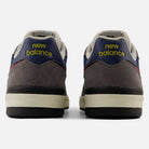 A rear view of the Castlerock Navy Jamie Foy Cup 306 CYZ New Balance Numeric Suede Skateboarding Shoes showcases their mainly grey suede upper with navy and red accents, highlighted by yellow "New Balance" text on the heel tabs against a light background. Inspired by pro skater Jamie Foy, these sneakers are crafted for impact protection while maintaining standout style.