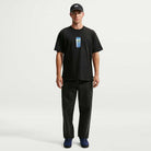 A man wearing a black cap, the Nike SB Skateboarding Cell Phone Tee Black IM4424010 T-shirt with a small chest graphic, loose black jeans, and dark sneakers stands facing forward against a plain white background.