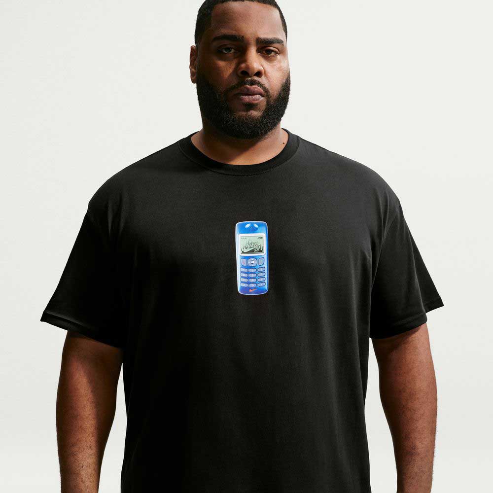 A man wearing the Nike SB Cell Phone Tee Black IM4424010 T-Shirt, featuring a blue retro cell phone graphic on the chest, stands against a plain white background, highlighting its sweat-wicking comfort and casual skateboarding style.