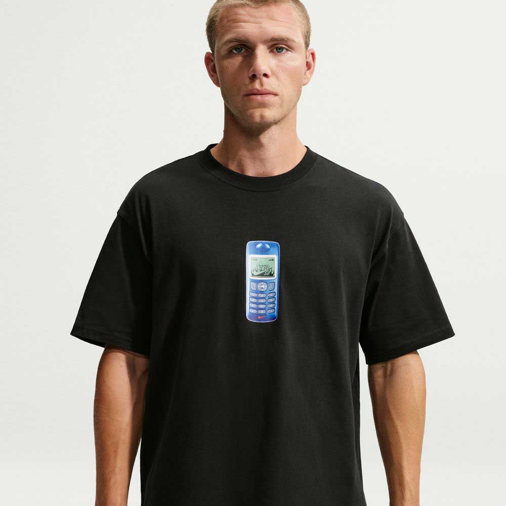 A man stands against a plain white background, wearing a black Nike SB Cell Phone Tee (IM4424010), featuring a graphic of a blue, old-fashioned mobile phone printed on the chest. Its sweat-wicking fabric offers comfort and complements the tee's effortless skate-inspired style.
