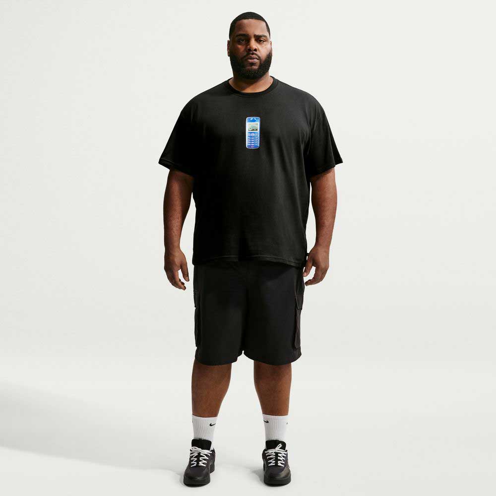 A man wearing a loose black Nike SB Cell Phone Tee Black IM4424010 T-Shirt, black shorts, white socks, and black sneakers stands facing forward against a plain white background. This sweat-wicking skate tee combines comfort and style for an effortlessly cool look.